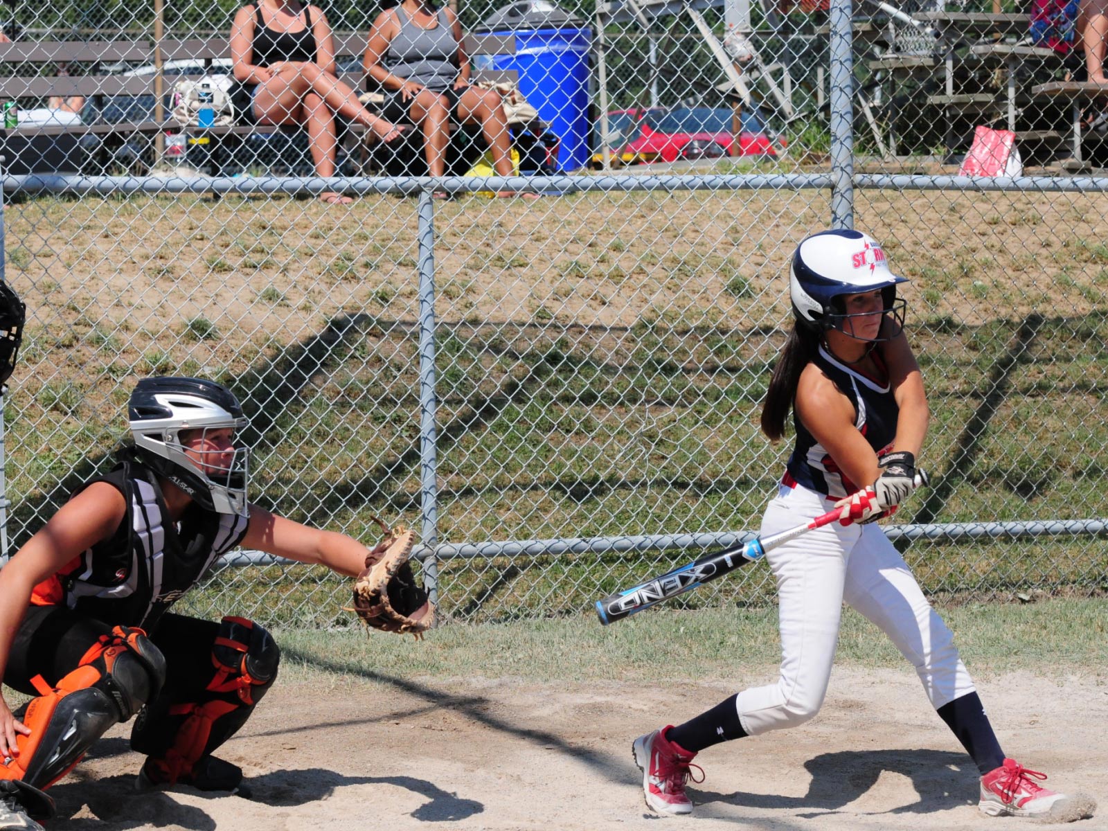 Young adult girls playing baseball on a baseball field. Patrons watching the game from behind the bleachers.